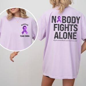 Pode incluir: Camiseta lil&aacute;s com a frase "NOBODY FIGHTS ALONE" nas costas e uma fita roxa. A frente apresenta uma fita roxa, a palavra "WARRIOR" e "TEAM EMMA".