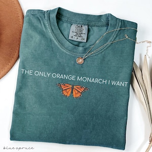 May include: A teal Comfort Colors t-shirt with the text "THE ONLY ORANGE MONARCH I WANT" and a graphic of an orange butterfly. A gold necklace with a pendant is draped on the shirt. A brown hat is visible in the upper left corner.