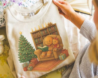 May include: Beige sweatshirt featuring a Christmas scene with a gingerbread person reading to a gingerbread girl in a cozy armchair. A Christmas tree, presents, and a bookshelf are also depicted. The sweatshirt is made of a soft material.