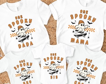 May include: White t-shirts and a onesie with a Halloween theme. Each shirt features the text "ONE SPOOKY" above a ghost riding a skateboard, with the words "DADA", "MAMA", "BRO", "SIS", and "CREW" below. Orange and black accents.