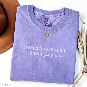 May include: A violet Comfort Colors t-shirt with the text "i teach tiny humans about jesus" in white script. A gold necklace with a pendant rests on the shirt. A brown hat is partially visible.
