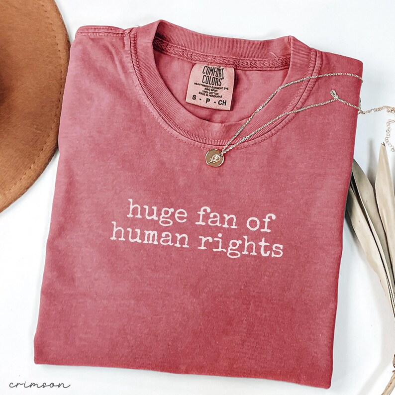 May include: Crimson-colored t-shirt with the text "huge fan of human rights" in white. A silver necklace with a small pendant rests on the shirt. The shirt has a Comfort Colors tag.