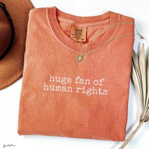 May include: A coral-colored t-shirt with the phrase "huge fan of human rights" printed in white. A gold necklace with a pendant rests on the shirt. A brown hat and dried plant are in the background.