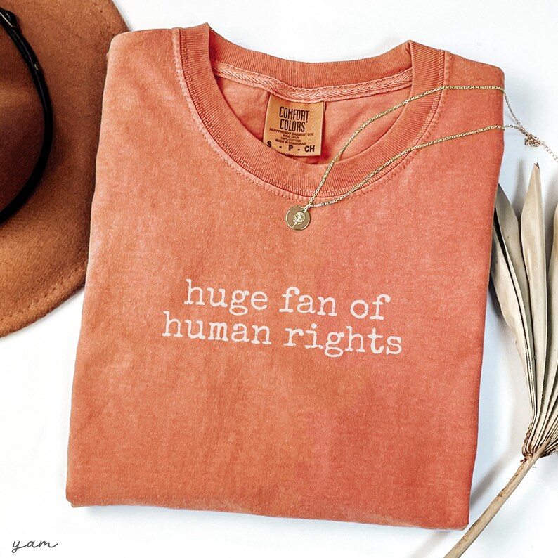 May include: A coral-colored t-shirt with the phrase "huge fan of human rights" printed in white. A gold necklace with a pendant rests on the shirt. A brown hat and dried plant are in the background.