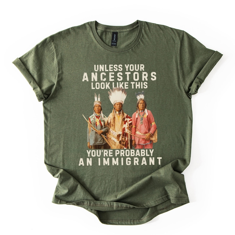 May include: Olive green t-shirt with the text "UNLESS YOUR ANCESTORS LOOK LIKE THIS YOU'RE PROBABLY AN IMMIGRANT." The graphic features three Native American figures in traditional attire. The shirt is made of a soft material.