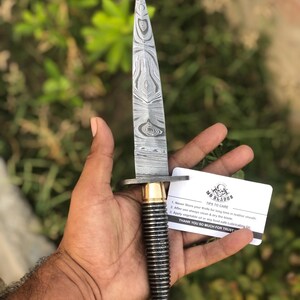 May include: A Damascus steel dagger with a patterned blade and a black and gold handle. The knife has a guard and a pointed tip. A card with care instructions is held near the knife.