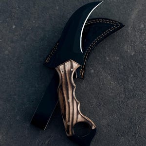 May include: A black karambit knife with a curved blade and a wooden handle with a finger hole. The knife is partially sheathed in a black leather case with white stitching. The blade has a white edge.