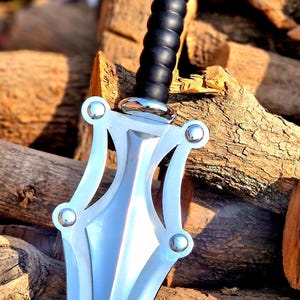 May include: A silver sword with a black handle and a decorative guard. The sword is resting on a pile of brown logs. The blade has a pointed tip and a curved guard with silver accents.