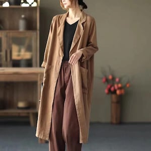May include: A long, tan linen coat with a notched collar and large front pockets. The coat is worn over a black top and brown pants. The outfit is a casual, yet stylish ensemble.