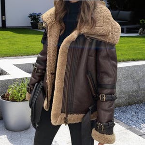 Puede incluir: Una chaqueta de aviador de cuero marrón y piel de oveja con cuello de piel y múltiples hebillas. La chaqueta tiene una cremallera y un forro de piel de oveja marrón.