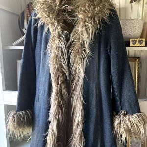 Könnte beinhalten: Ein langer, dunkelblauer Jeansmantel mit einem Kunstfellkragen, -besatz und -manschetten. Das Kunstfell ist hellbraun. Der Mantel hängt an einem Kleiderbügel.