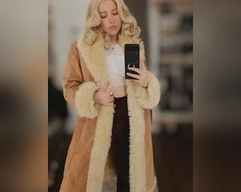 May include: A long, tan suede coat with cream-colored faux fur trim on the collar, cuffs, and along the front edges. The coat is knee-length and has a vintage, retro style. The model is wearing the coat.