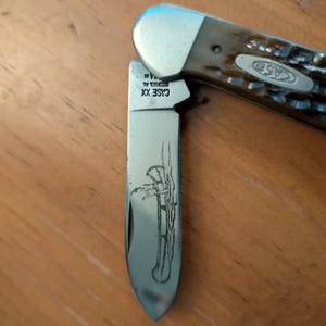 May include: A folding pocket knife with a brown textured handle and a silver metal bolster. The blade features an etched illustration of a person paddling a canoe and the text CASE XX. The knife is partially open, revealing the metallic blade.