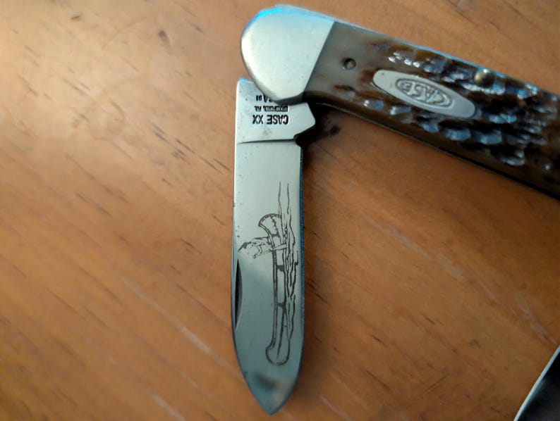 May include: A folding pocket knife with a brown textured handle and a silver metal bolster. The blade features an etched illustration of a person paddling a canoe and the text CASE XX. The knife is partially open, revealing the metallic blade.