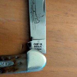 May include: A folding pocket knife with a metallic blade featuring an etched canoe design. The tang stamp reads CASE XX BRADFORD PA USA 91. The handle is made of a textured brown material with a silver bolster and a small inlaid CASE logo emblem.