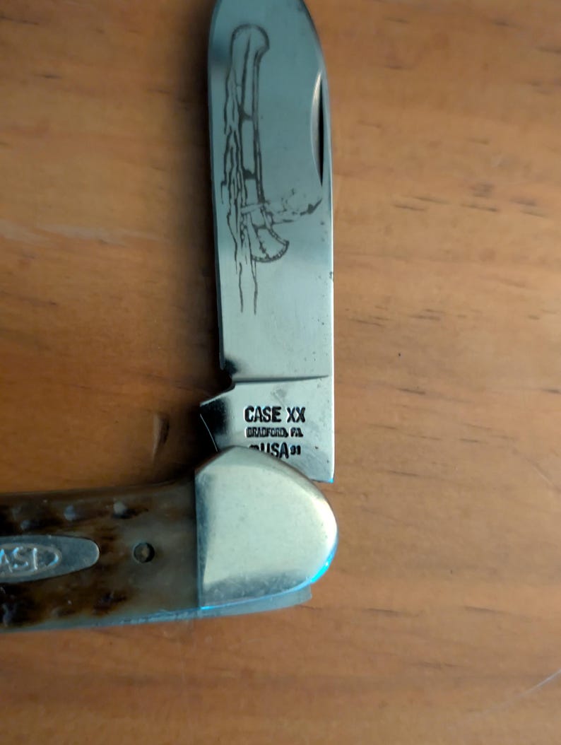 May include: A folding pocket knife with a metallic blade featuring an etched canoe design. The tang stamp reads CASE XX BRADFORD PA USA 91. The handle is made of a textured brown material with a silver bolster and a small inlaid CASE logo emblem.