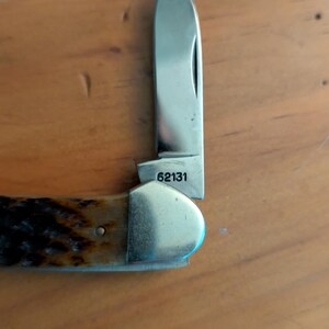 May include: A folding pocket knife with a single silver-colored steel blade extended upward. The base of the blade is clearly stamped with the number 62131. The handle features a textured, dark brown, and tan mottled pattern with a polished metal bolster.