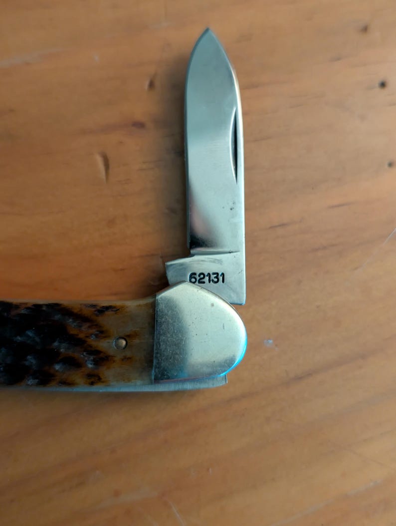 May include: A folding pocket knife with a single silver-colored steel blade extended upward. The base of the blade is clearly stamped with the number 62131. The handle features a textured, dark brown, and tan mottled pattern with a polished metal bolster.