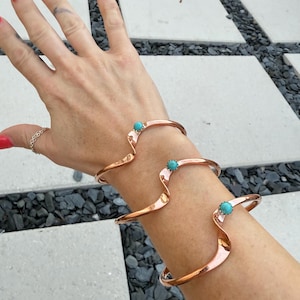 May include: Three copper-colored, open-ended bracelets with a wavy design, each featuring a small, round turquoise stone. The bracelets are worn on a wrist, showcasing their unique shape and color.