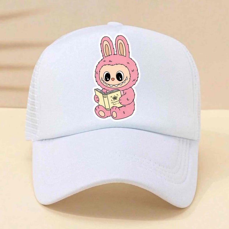 May include: White baseball cap with a mesh back and a curved brim. The cap features a cartoon illustration of a pink bunny character reading a book. The bunny has large eyes and is wearing a pink outfit. The book has a floral design.
