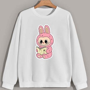 May include: White sweatshirt featuring a pink cartoon bunny character reading a book. The bunny is wearing a pink costume with large ears and a fluffy texture. The book is yellow with a floral design.