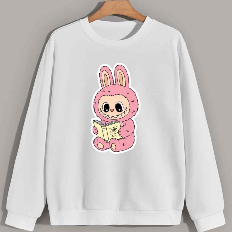 May include: White sweatshirt featuring a pink cartoon bunny character reading a book. The bunny is wearing a pink costume with large ears and a fluffy texture. The book is yellow with a floral design.