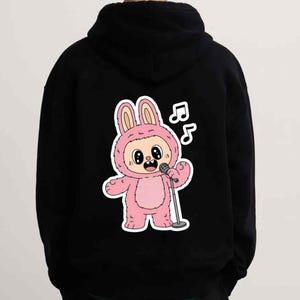 May include: Black hoodie featuring a cartoon bunny character in a pink costume holding a microphone. The bunny has large eyes and is singing, with musical notes above its head. The hoodie has a drawstring hood.