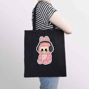 May include: Black tote bag featuring a pink cartoon character wearing headphones. The character has a bunny-like appearance with large eyes and a smiling mouth. The bag is held by a person wearing a black and white striped shirt.