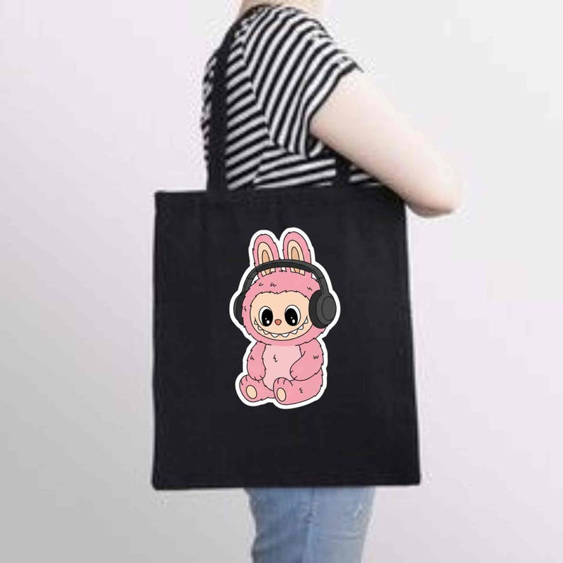 May include: Black tote bag featuring a pink cartoon character wearing headphones. The character has a bunny-like appearance with large eyes and a smiling mouth. The bag is held by a person wearing a black and white striped shirt.