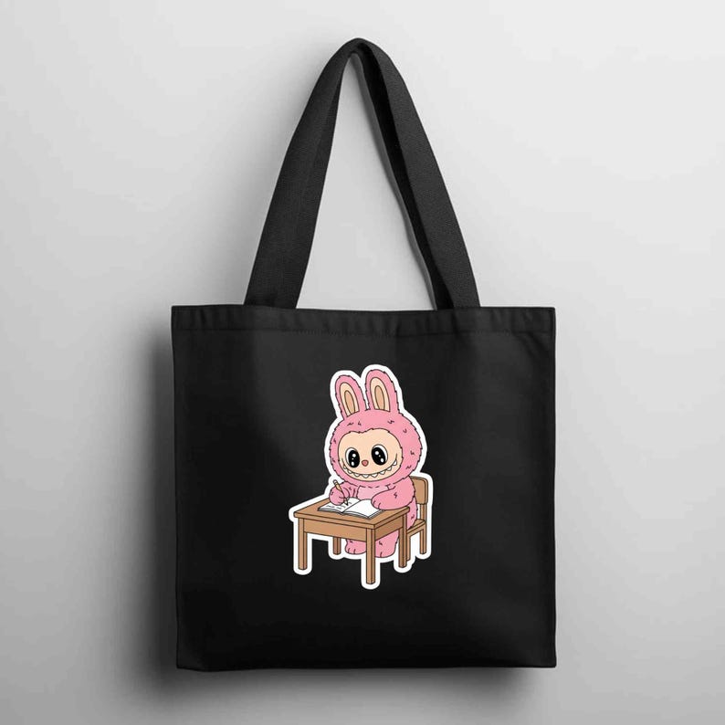 May include: Black tote bag featuring a pink bunny character in a costume, seated at a small wooden desk and writing. The bunny has a smiling face and is wearing a pink hooded outfit. The bag has two black handles.