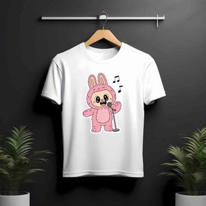 May include: White t-shirt featuring a cartoon character in a pink bunny costume singing into a microphone. The bunny has large eyes and is holding a silver microphone. Musical notes are above the bunny. The shirt is hanging on a wooden hanger.