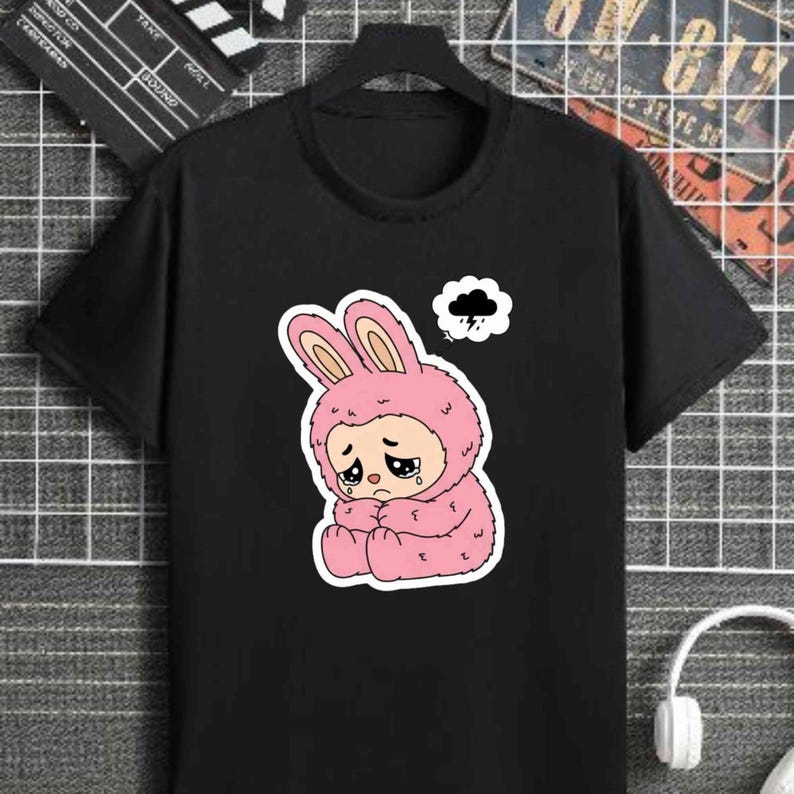 May include: Black t-shirt featuring a pink cartoon bunny character with a sad expression and tears. The bunny is wearing a hooded costume and a thought bubble with a rain cloud above its head. The shirt has short sleeves.