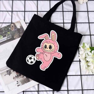 May include: Black tote bag featuring a pink cartoon bunny in a soccer uniform kicking a soccer ball. The bunny has a smiling face and is wearing a bunny suit. The bag has two black handles.