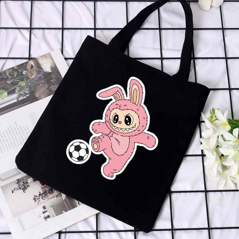 May include: Black tote bag featuring a pink cartoon bunny in a soccer uniform kicking a soccer ball. The bunny has a smiling face and is wearing a bunny suit. The bag has two black handles.