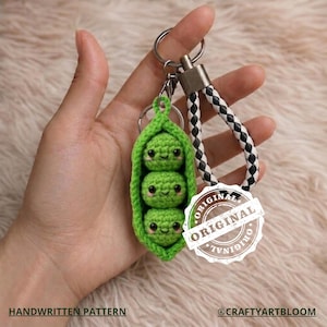 Peut inclure: Un porte-clés fait main en forme de cosse de pois verte au crochet, avec trois petits pois souriants. Le porte-clés est doté d'un anneau argenté et d'une lanière tressée noire et blanche. Le texte "HANDWRITTEN PATTERN" et "@CRAFTYARTBLOOM" est également visible.