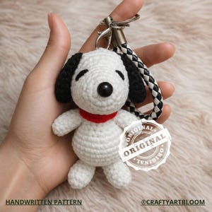 May include: Crocheted Snoopy keychain with black ears, a black nose, and a red collar. The white body is accented with black stitching. The keychain has a silver clasp and a black and white braided strap. The text "HANDWRITTEN PATTERN" and "ORIGINAL" are visible.