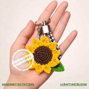 May include: A handmade sunflower keychain with a yellow crocheted flower and a brown center. The keychain has a silver metal ring and a black and white braided strap. The tag reads "ORIGINAL". The text "HANDWRITTEN PATTERN" is at the bottom.