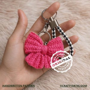 May include: A handmade pink crocheted bow keychain with a braided black and white strap. The keychain has a silver ring and clasp. The text "ORIGINAL" is stamped on the image. The text "HANDWRITTEN PATTERN" and "@CRAFTYARTBLOOM" are also visible.