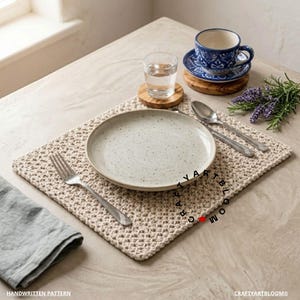 Puede incluir: Un mantel individual rectangular de ganchillo beige con un patrón de puntos texturizado. Centrado sobre el mantel hay un plato de cerámica blanquecino moteado, un tenedor de plata, una cuchara de plata y una taza azul y blanca.