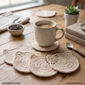 Könnte beinhalten: Ein Set aus beigen, katzenförmigen Häkeluntersetzern aus Garn. Ein Untersetzer liegt unter einer gesprenkelten Keramiktasse mit Kaffee. Die Untersetzer haben abgerundete Ohren und eine kreisförmige Basis. Der Text CRAFTYARTBLOOM ist auf den Untersetzern zu sehen.