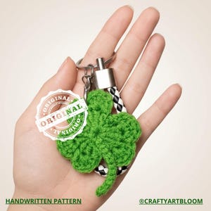 Può includere: Un portachiavi a forma di quadrifoglio verde all'uncinetto con un portachiavi argentato e un cinturino intrecciato bianco e nero. Il portachiavi ha un timbro "ORIGINAL" e il testo "HANDWRITTEN PATTERN".