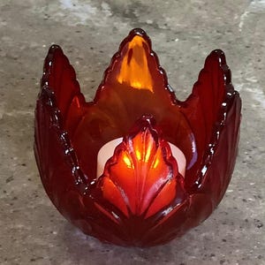 May include: A red glass candle holder shaped like a stylized flower or leaf. The candle holder has a glossy finish and a white candle inside, with the flame casting an orange glow.