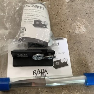 May include: A black RADA Cutlery knife sharpener with two sharpening wheels, a clear tube with a blue cap, and product packaging. The packaging includes the text "Knife sharpener" and "Lifetime Guarantee".