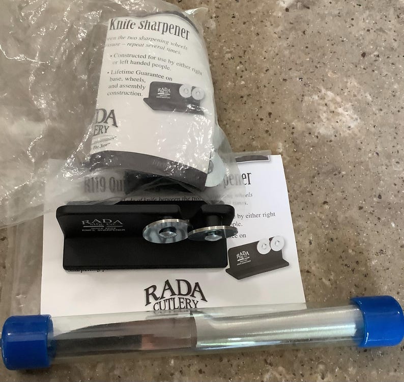 May include: A black RADA Cutlery knife sharpener with two sharpening wheels, a clear tube with a blue cap, and product packaging. The packaging includes the text "Knife sharpener" and "Lifetime Guarantee".