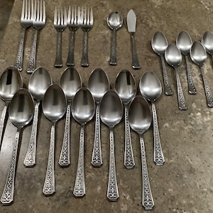 May include: A collection of silver-colored flatware, including forks, spoons, and a butter knife, arranged on a neutral surface. The utensils feature ornate, detailed handle designs. The set includes various sizes of spoons and forks, suitable for a formal dining setting.