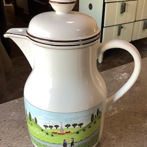 May include: White ceramic coffee pot with a lid and handle. The pot features a brown trim and a landscape scene with figures, trees, and a church. The pot is likely vintage.