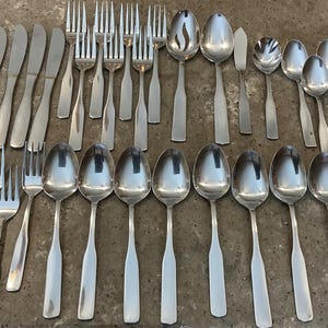 May include: A large collection of silver-toned stainless steel flatware including dinner knives, forks, and various sizes of spoons. The set features a simple, smooth handle design with rounded ends, arranged in rows on a flat surface.