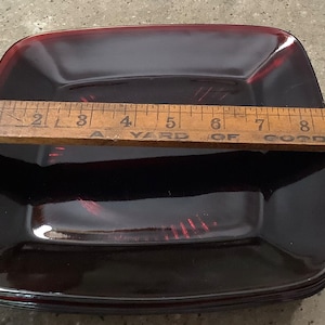 May include: A stack of four square, ruby-red glass plates. A wooden ruler rests across the plates, measuring approximately 9 inches. The plates have a slightly raised edge and a glossy finish, suitable for serving food.