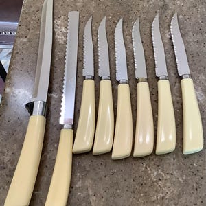 May include: A set of eight kitchen knives featuring smooth cream-colored handles and silver-toned metal blades. The collection includes one long straight-edged carving knife, one long serrated bread knife, and six smaller serrated steak knives.