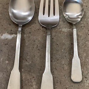 May include: A set of three silver-colored utensils: a spoon, a fork, and a smaller spoon. The spoon has an oval bowl, the fork has four tines, and all pieces have rectangular handles. The utensils are arranged on a speckled surface.